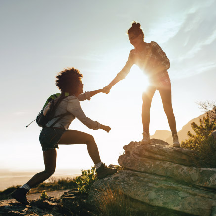 Two women hiking. With hands together, one woman is helping the other step up onto a rocky surface.