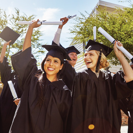 Group of young adults wearing graduation caps and gowns, holding diplomas up in air.