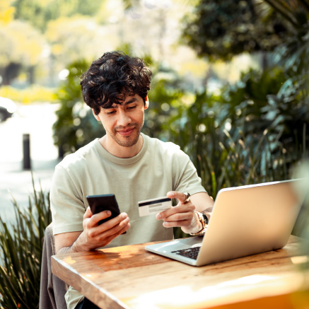 A man sitting on a patio holding a phone, debit/credit card in front of a laptop