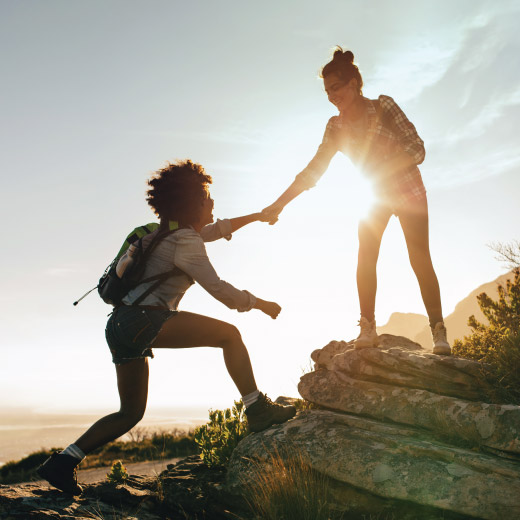 Two women hiking. With hands together, one woman is helping the other step up onto a rocky surface.